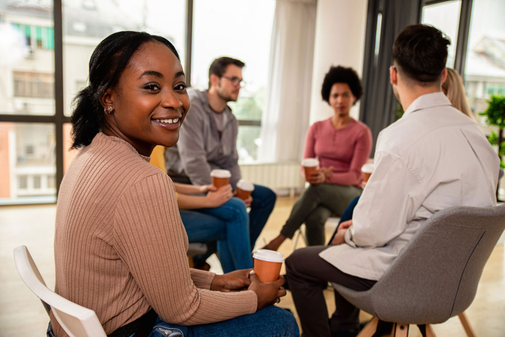 A woman smiles at the camera while sitting with a group of people in a circle, all holding disposable coffee cups, in a bright room with large windows.