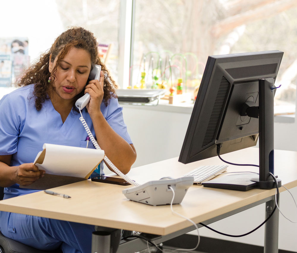 A woman in blue scrubs sits at a desk, talking on a phone while reading from a clipboard. A computer monitor and phone are on the desk, with a window and toys visible in the background.