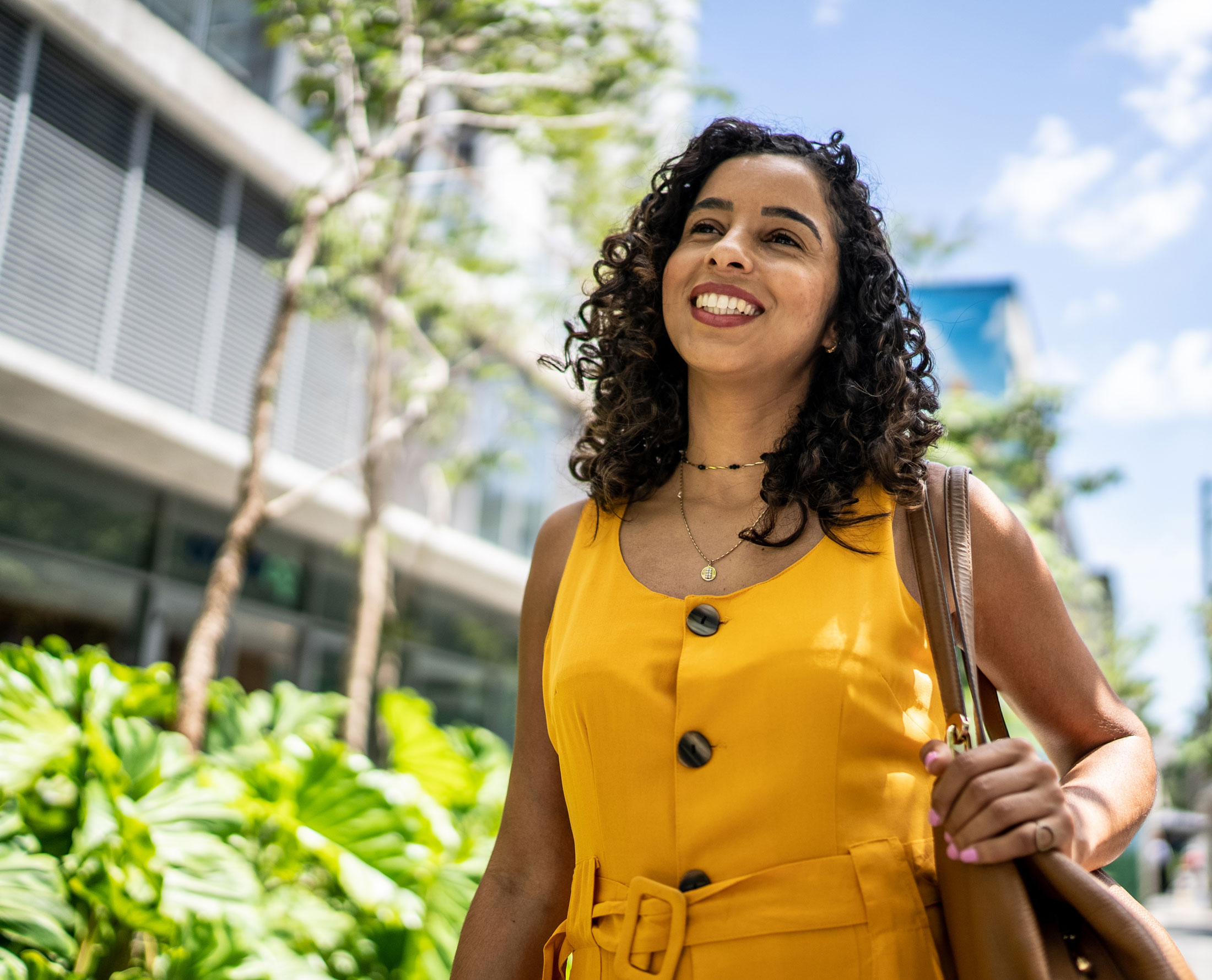 A woman with curly hair, wearing a yellow sleeveless dress and carrying a brown bag, smiles while walking outdoors on a sunny day with green plants and modern buildings in the background.