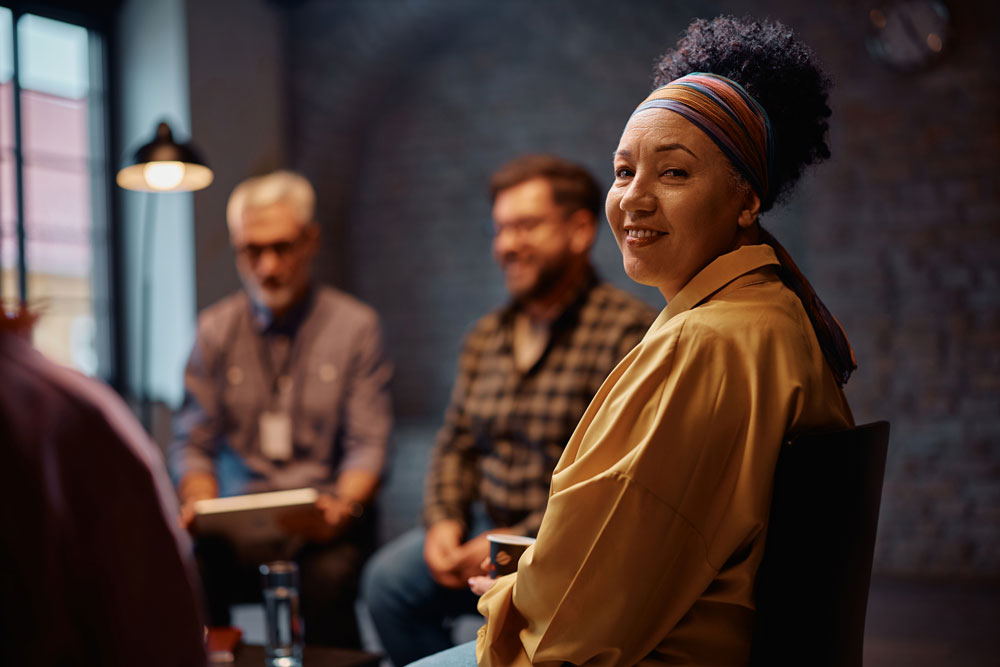 A woman in a yellow shirt and colorful headband smiles at the camera while sitting in a group therapy or support session, with two people talking and blurred in the background.