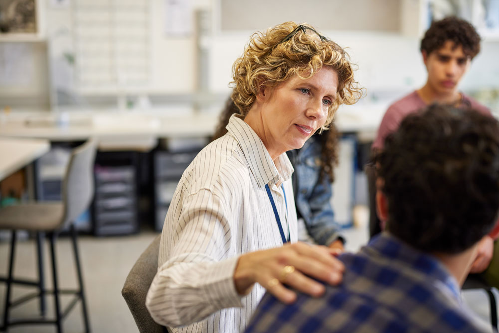 A woman with curly hair wearing a striped shirt sits in a classroom, gently placing her hand on a student’s shoulder while listening attentively. Other students are seated nearby, engaged in the discussion.