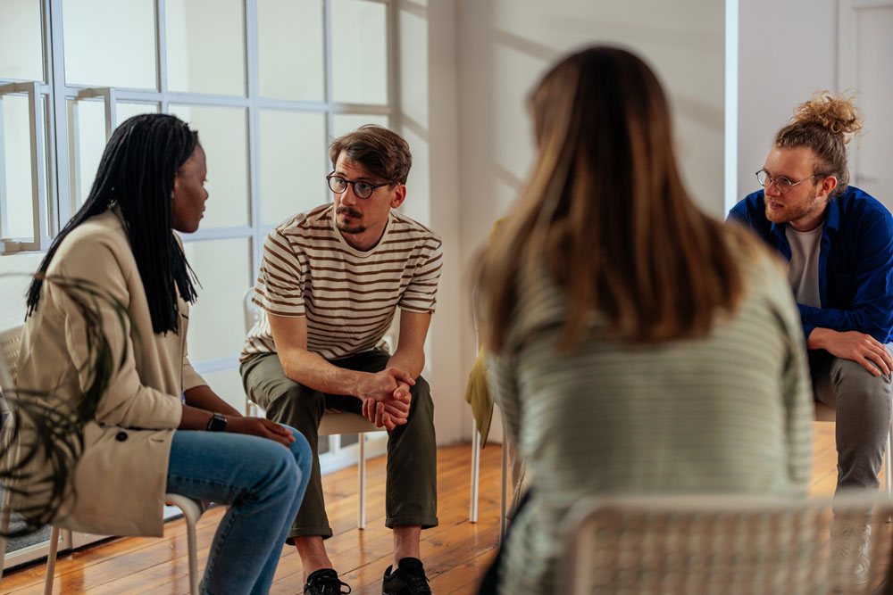 Four people sit in a circle indoors, engaged in a serious conversation. One person speaks while the others listen attentively. The setting appears informal and supportive, possibly a group discussion or therapy session.