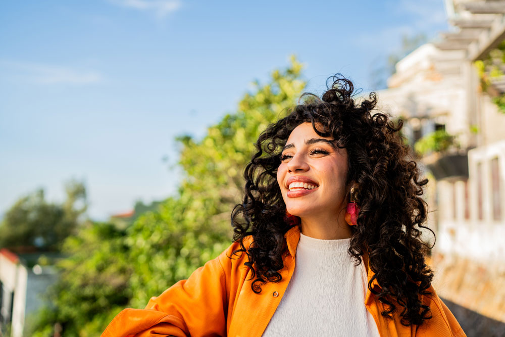 A woman with curly hair wearing a bright orange jacket and white top smiles joyfully outdoors, with greenery and blue sky in the background on a sunny day.