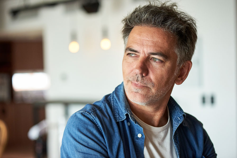 A middle-aged man with gray hair and stubble, wearing a blue denim shirt over a white t-shirt, looks thoughtfully to the side in a softly lit indoor setting with blurred lights in the background.