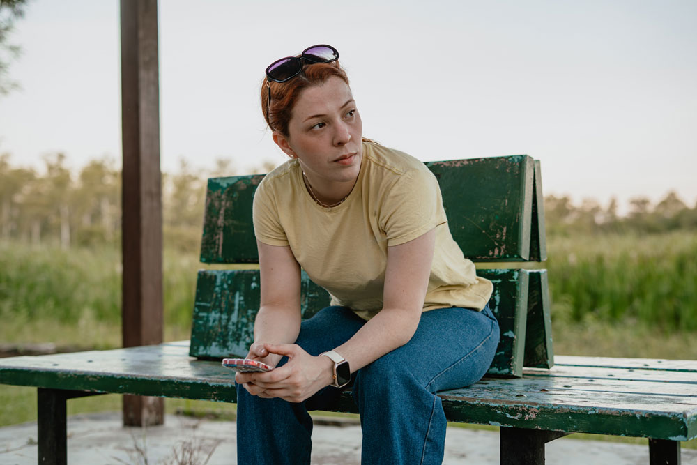 A person with short red hair, wearing a yellow t-shirt, blue jeans, and sunglasses on their head, sits on a weathered green bench outdoors, holding a phone and looking to the side.